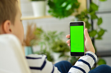 The boy dressed in a striped longsleeve sits in a white armchair and is holding a phone in his hand with a green screen.