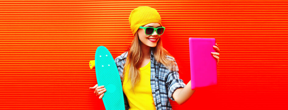 Portrait of stylish cool young woman using tablet pc with skateboard wearing colorful clothes on vivid background