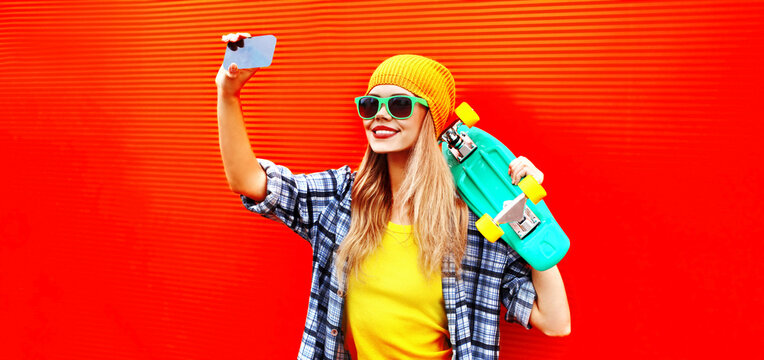 Summer portrait of happy smiling young woman model taking selfie by smartphone with skateboard on colorful orange background