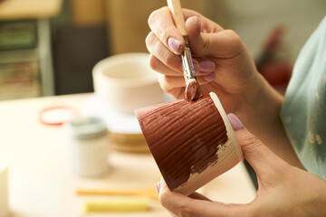 Close-up of girl painting clay mug with glaze. Woman coloring pottery in workshop with a paintbrush. Painter in green apron glazing clay pot. - 491521021