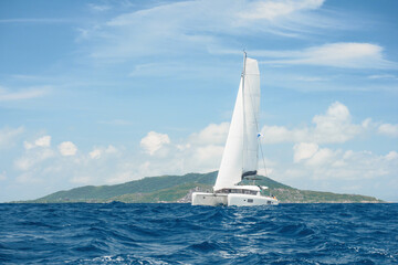 Luxurious white catamaran is sailing at full sail on the blue ocean water, moving away from the...