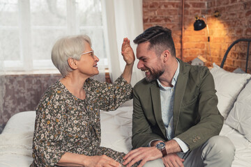 European grey-haired grandmother sitting on her industrial bed and comforting laughing her son. High quality photo