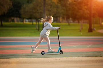 Portrait of active little toddler girl riding scooter on road in park outdoors on summer day....