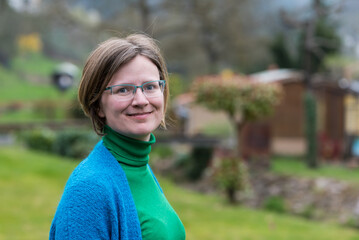 Close up portrait of a young white woman with glasses wearing a blue and green sweater with a green blurry nature background - obrazy, fototapety, plakaty