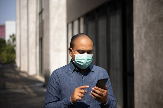 Young indian using smartphone standing in front of office building. Asian man wearing protective mask during pandemic. - Powered by Adobe