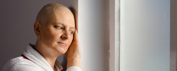 Young bald woman in bathrobe suffering from cancer looking through hospital window.