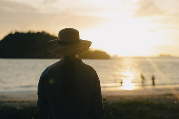 Young asian woman with straw hat standing alone on beach looking at sunset seashore. Chilling in holiday weekend summertime. Traveler Female walking around the beach with the sunlight