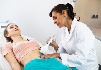 Confident beautician making needle-free mesotherapy procedure for young adult woman on belly for reducing silhouette imperfection