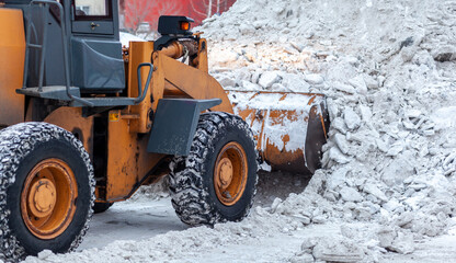 Big orange tractor cleans up snow from the road and loads it into the truck. Cleaning and cleaning of roads in the city from snow in winter. Snow removal after snowfall and blizzards. 