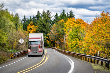 Fototapeta na wymiar Dark red classic powerful big rig semi truck transporting commercial cargo driving on the winding road through the autumn forest