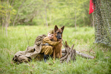 a boy in a military uniform in a clearing, sitting by a campfire with a German shepherd.Two friends...
