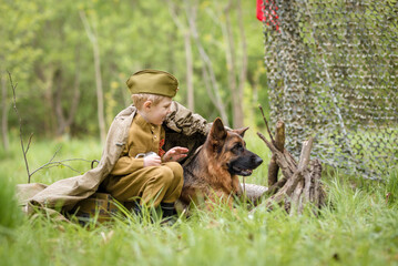 a boy in a military uniform in a clearing, sitting by a campfire with a German shepherd.Two friends defend the motherland