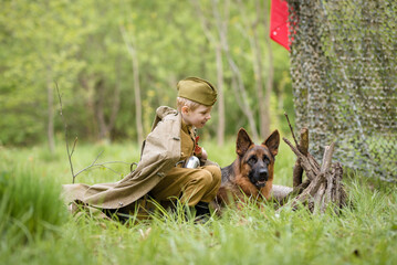 a boy in a military uniform in a clearing, sitting by a campfire with a German shepherd.Two friends...