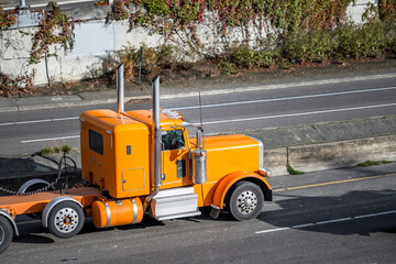Orange classic stylish big rig semi truck with extended frame transporting cargo driving on the divided highway road
