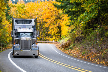 Powerful old style dark gray classic big rig semi truck with refrigerator unit on the front wall of the reefer semi trailer running on the winding mountain road through the autumn forest