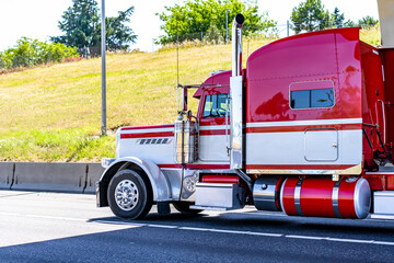 Shiny red big rig classic bonnet semi truck tractor with long truck driver rest compartment and chrome parts transporting cargo running on the sunny highway road