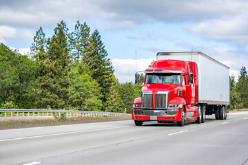 Stylish bright red big rig semi truck with chrome parts transporting cargo in dry van semi trailer running on the straight wide highway road with green trees on the side © vit