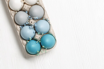 Colorful Easter eggs and flowers decor in carton pack, blue, beige, white neutral colors, festive chicken egg in paper container on white wooden table, decorated holiday food