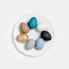 Colorful Easter eggs on round white plate, shine golden, blue, gray, beige, black, natural colored. Dyed Chicken egg various colors. Easter holiday food top view, minimal design aesthetic