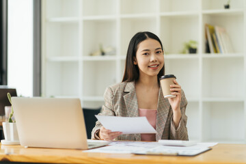 Asian beautiful young business woman drinking coffee working on desk with laptop and financial report document.have break time and resting after solving task.
