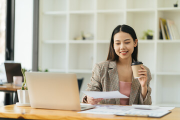 Asian beautiful young business woman drinking coffee working on desk with laptop and financial report document.have break time and resting after solving task.