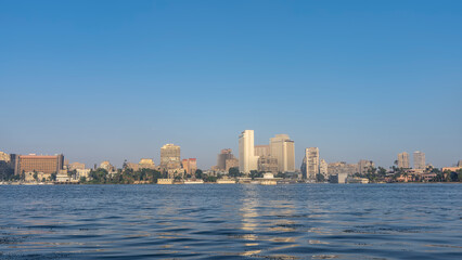 Obraz premium On the banks of the Nile in Cairo, city houses and green vegetation are visible. Ripples and reflections on the surface of blue water. Clear azure sky. Egypt.