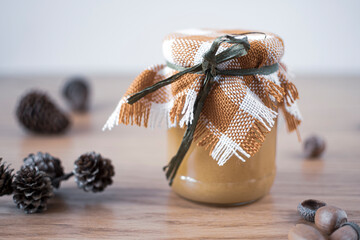 Decorative jar with honey and pine cones on a table. Natural seamless fabric on a glass container for sweet organic liquid. Selective focus on the details, blurred background.