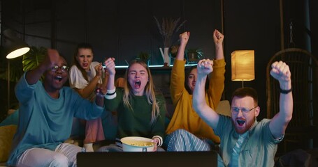 Happy young multiethnic sports fans sit together watching football on TV at home with snacks celebrating goal and win.