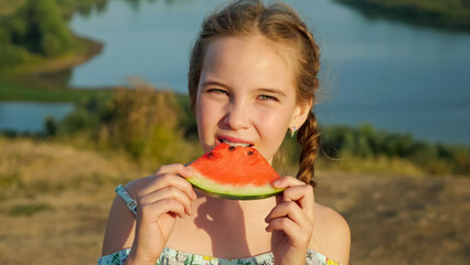 Pretty young schoolgirl with blonde plait bites slice of delicious juicy watermelon standing on riverbank on sunny day extreme close view