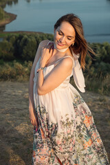 Seductive brunette woman in loose dress with flower pattern and scarf poses for camera on tranquil...