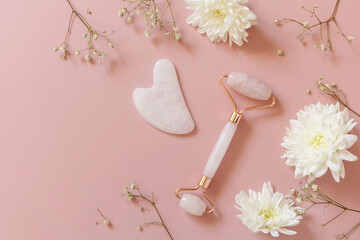 Rose quartz facial massage roller over pink background with gypsophila flowers. Massage tools with jade stone, anti-aging, anti-wrinkle beauty skincare tool. Top view flat lay. Copy space. - obrazy, fototapety, plakaty