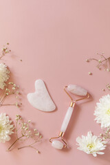 Rose quartz facial massage roller over pink background with gypsophila flowers. Massage tools with jade stone, anti-aging, anti-wrinkle beauty skincare tool. Top view flat lay. Copy space.