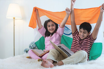 Indian brothers and sisters with laptops on lap sitting on bed, holding Indian flags together,...