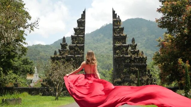 Pretty asian woman walks to balinese gates in long red dress in slowmotion on Hangara gates in Bali Indonesia. High quality fullhd footage