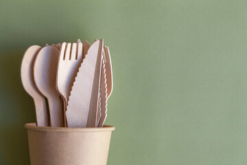 Wooden disposable cutlery (forks, spoons and knifes) lies in brown paper coffee cup on green...