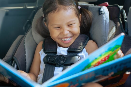A little kid sitting in her car seat, reading a book smiling.