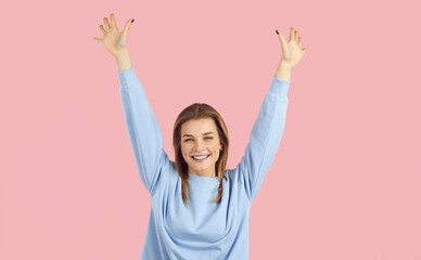 Studio shot of a happy joyful young woman raising her arms and smiling. Cheerful beautiful girl in a pale blue sweatshirt standing with her hands up isolated on a solid pastel pink background