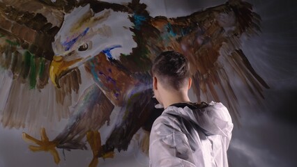 Fototapeta na wymiar Artist designer draws an eagle on the wall. Craftsman decorator paints a picture with acrylic oil color. Painter painter dressed in a paint coat. Indoor. Dark magic cinematic look.