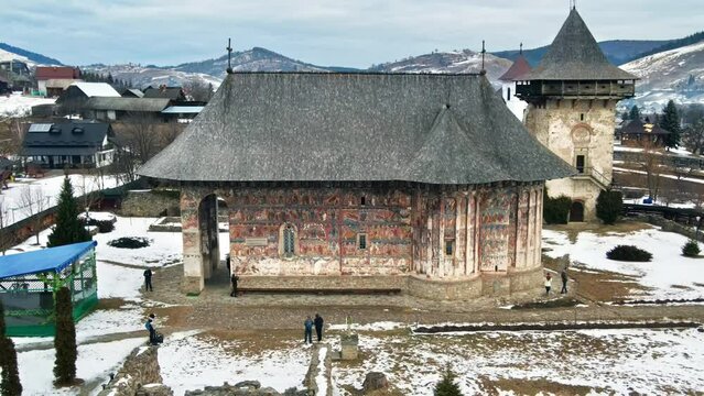 Aerial drone view of The Humor Monastery in winter, Romania. Main painted church and the Tower, yard with snow and tourists, bare trees, residential buildings