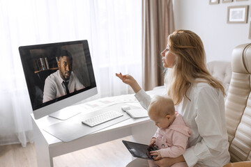 Working young mother in maternity leave work remotely or study at home on computer while infant daughter hold papers and looking at screen. Modern day mother of little child sit at desk use gadgets