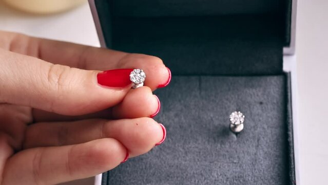 Close view of a woman holding earring with diamonds. A box with another earring behind, hands with red nails