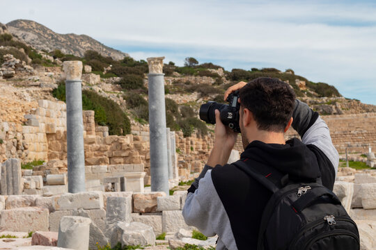 Back view of young traveler tourist boy. He is taking photograph of historical columns  in Knidos ancient city. It was a Greek city in ancient Caria, located by sea Datca, Mugla, Turkey. 