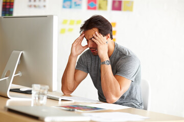 Exhaustion kills creativity. An overwhelmed man sitting at an office desk with his head rested against hands.