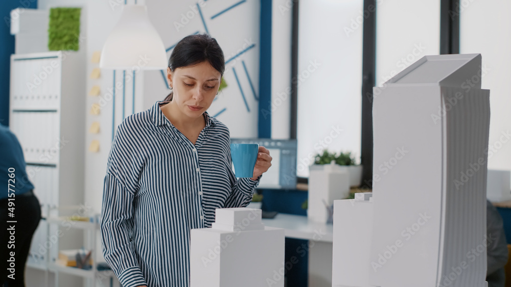 Poster portrait of woman engineer looking at building model and drinking cup of coffee at work. architect a