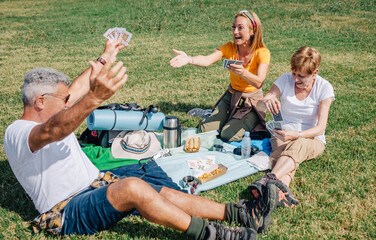Senior man celebrating that he has won his family playing cards during an excursion sitting on a blanket outdoors