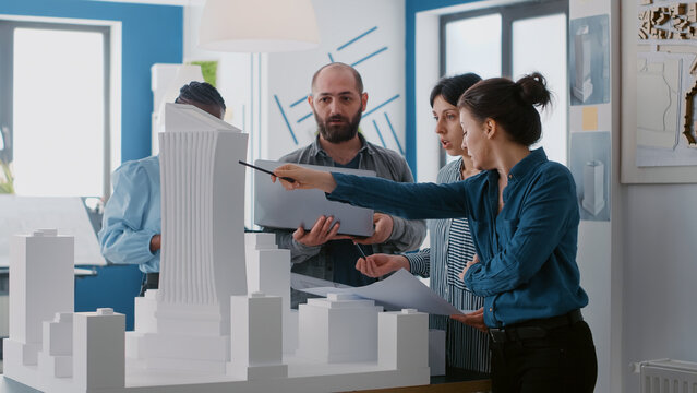 Multi ethnic people working with laptop, tablet and building model for architecture project. Coworkers doing teamwork to design maquette and construction layout with blueprints.