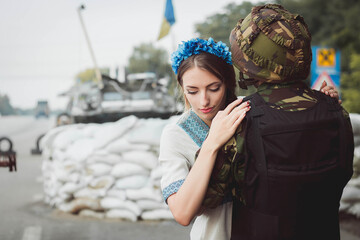 Ukrainian soldier embraces young woman on roadblock against background of sandbags.