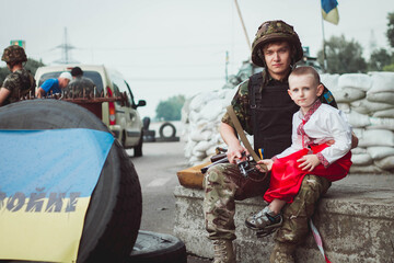 Ukrainian child boy sits near ukrainian soldier on roadblock against background of sandbags.
