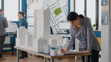 Woman architect analyzing blueprints plan on table to design building model and maquette. Engineer working on architectural development project with industrial construction tools.