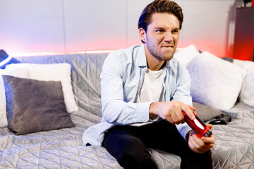 Concentrated guy playing video game, holding remote red controller, enjoying leisure time with device, engaged in online competition with friends,having fun at home during quarantine,sitting on couch